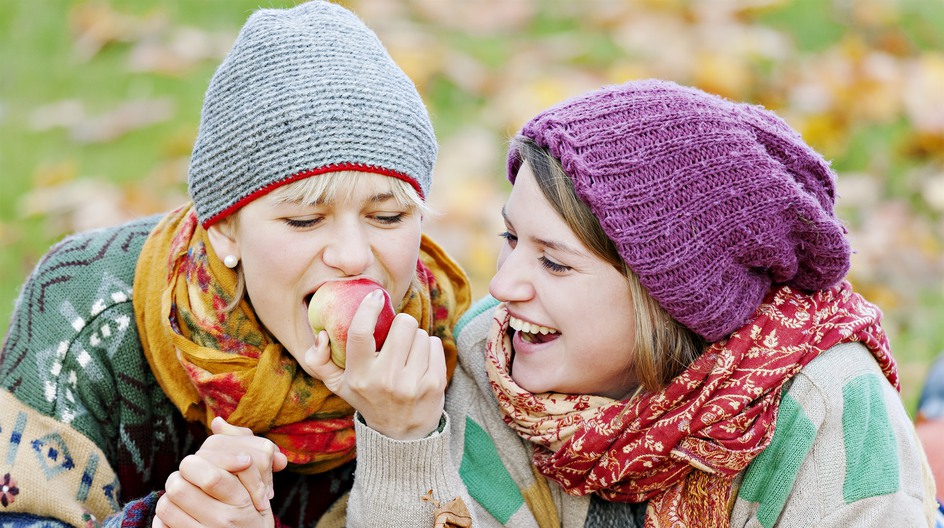 Två kvinnor iförda mössor delar på ett äpple i höstlöven